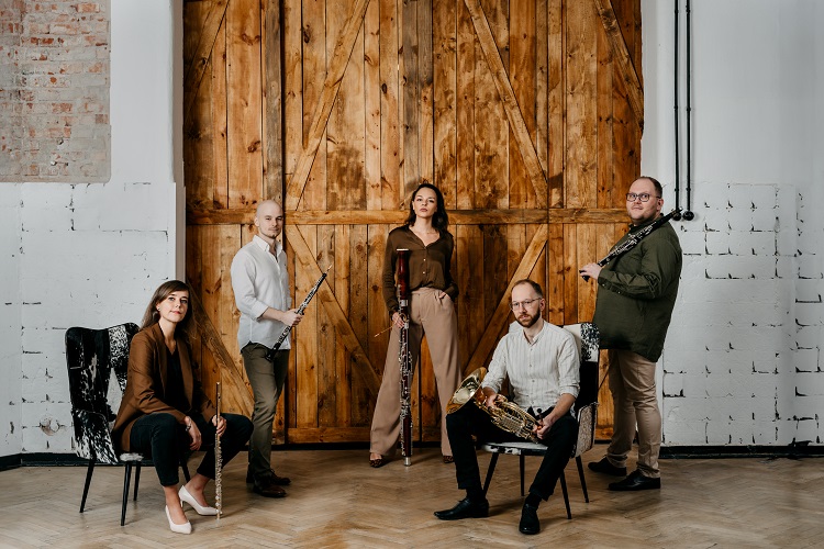 Pięcioosobowa grupa muzyków stoi na tle drewnianych drzwi w industrialnym wnętrzu. Każdy trzyma instrument: klarnet, flet, fagot, waltornia, obój. Dwie osoby siedzą na czarnych krzesłach po bokach, pozostałe stoją pośrodku, prezentując poważne i skupione wyrazy twarzy. Cracow Golden Quintet, fot. Anita Wąsik