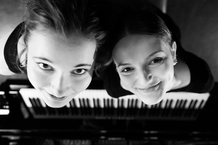 Czarno-białe zdjęcie dwóch młodych kobiet stojących przy fortepianie, sfotografowanych od góry. Patrzą w obiektyw z uśmiechem. Reverie Piano Duo, fot. Adam Kujawski