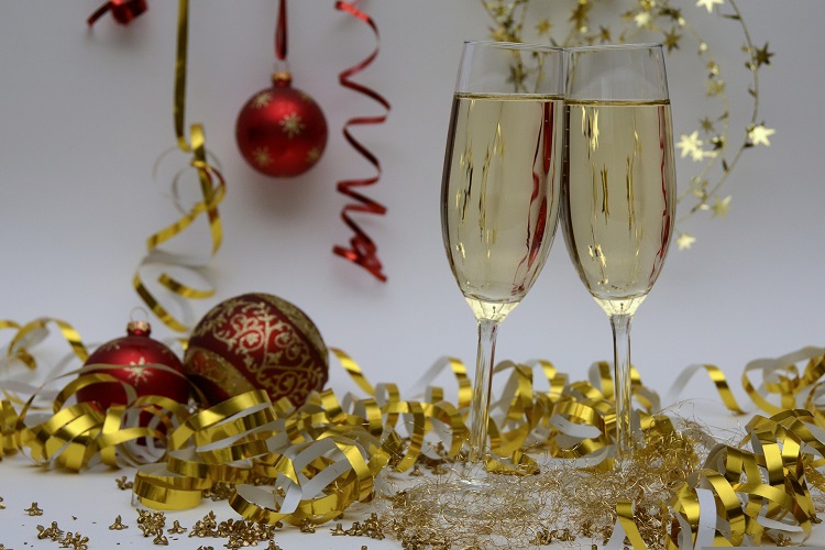 Eleganckie kieliszki wypełnione szampanem, umieszczone na tle rozrzuconych złotych i czerwonych serpentyn. Wiszące bombki choinkowe dodają świątecznego charakteru kompozycji.